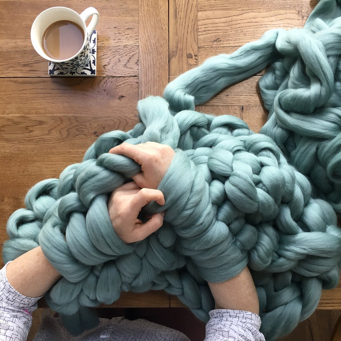 Arms giant knitting a light teal blanket with a cup of tea to the side 